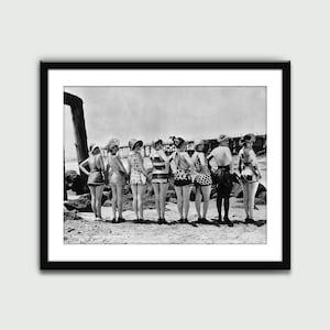 Könnte beinhalten: Ein Schwarzweißfoto von acht Personen, die auf einem Strand stehen und der Kamera den Rücken zukehren. Sie tragen alle Badeanzüge und Hüte. Die Personen stehen in einer Reihe, wobei die Person links am nächsten zur Kamera steht. Der Strand ist sandig und im Hintergrund befindet sich ein Steg.