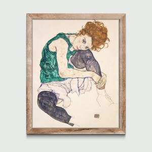 May include: Framed artwork featuring a seated figure with red hair, wearing a green top and dark pants. The artwork is a sketch with a light-colored background and a wooden frame. The figure is in a seated position, with their legs drawn up.