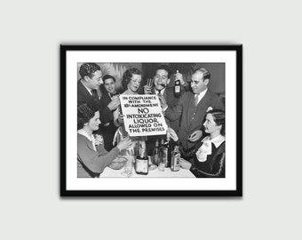 May include: A black and white photo of a group of people gathered around a table, holding bottles and glasses. They are holding a sign that reads "In compliance with the 18th Amendment, No intoxicating liquor allowed on the premises."