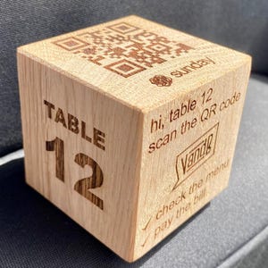 May include: A wooden cube with engraved text and a QR code. The cube reads "TABLE 12" on one side and "hi, table 12 scan the QR code" on another. The top of the cube has a QR code and the word "sunday".