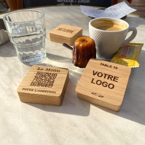 Puede incluir: Cuatro posavasos de madera con texto grabado. Los posavasos están sobre una mesa de mármol con un vaso de agua, una taza de café y un pastel. El texto en los posavasos dice "Reserve", "Le Menu", "Mesa 10" y "Su Logo".