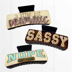 May include: Three rectangular hair claw clips with black clasps. One clip reads "a little bit Dramatic" in pink glitter, another says "SASSY" in a checkered pattern, and the last says "NOPE NOT TODAY" in turquoise.