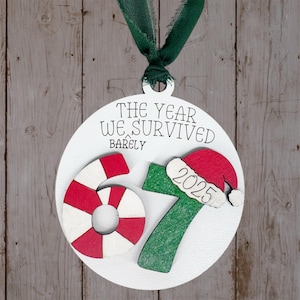 May include: A white Christmas ornament with the text "THE YEAR WE SURVIVED BARELY". It features a red and white candy cane, a green number 7, and a Santa hat with "2025". A green ribbon is attached for hanging.