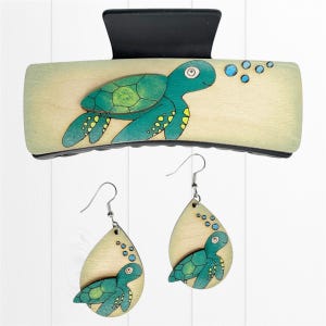 May include: A wooden hair clip and matching earrings set. The hair clip is rectangular with a black clasp and a painted teal sea turtle design. The earrings are teardrop-shaped with a sea turtle design and silver hooks. The sea turtles have yellow accents.