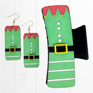 May include: A set of Christmas-themed accessories, including a pair of earrings and a hair clip. The earrings and clip are designed to resemble an elf's outfit, featuring green, red, and yellow accents. The earrings have gold-colored hooks.