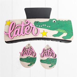 May include: A black hair claw clip and two matching teardrop-shaped earrings. Each piece features a light wood base with a green alligator illustration, the word later in pink script, and small yellow stars. The set displays a cohesive design.