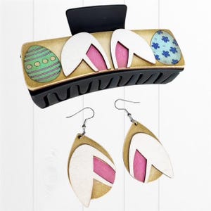 May include: A set of Easter-themed hair accessories featuring a brown and black hair clip with bunny ears and two Easter eggs, and a pair of dangle earrings with bunny ears and a pink and white design.