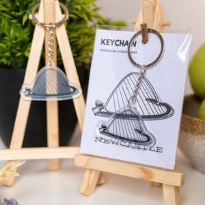 Puede incluir: Una tarjeta blanca con el texto "KEYCHAIN DESIGNED BY @THEMODBOD" y "NEWCASTLE" en ella. Un llavero plateado con un dije de acrílico transparente del Puente Tyne en Newcastle upon Tyne, Inglaterra, se muestra en la tarjeta. El llavero está en un pequeño caballete de madera.