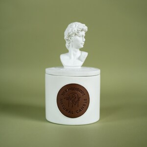 Op de afbeelding: Een witte keramische kaars pot met een gesculpteerd buste van een man erop. De pot heeft een bruin lederen label met de tekst "The MediBody Natural Candle".