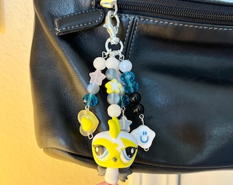 May include: A yellow and white bird charm with pink feet and a blue eye. The charm is attached to a silver chain with blue, white, and yellow beads, including stars and a smiley face charm.