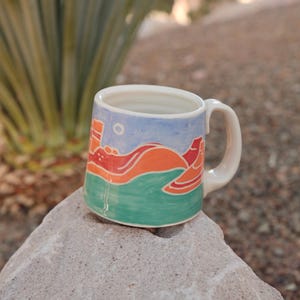 Peut inclure: Une tasse en céramique avec un paysage. La tasse présente un ciel bleu, des collines orange et rouges et de l'eau verte. La poignée et l'intérieur sont blancs. La tasse est posée sur un rocher gris.