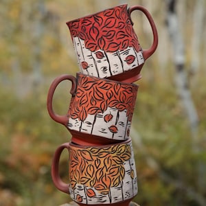Peut inclure: Une pile de trois tasses en céramique avec une base couleur terre cuite. Chaque tasse présente un motif sculpté de troncs de bouleaux blancs avec des marques noires et un feuillage dans des tons de rouge, orange et jaune, avec une anse.