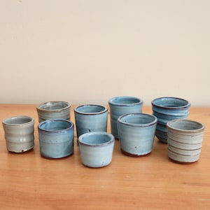 May include: A collection of nine small ceramic cups in various shades of blue and grey. Each cup features a unique cylindrical shape with subtle variations in height, width, and surface texture, including smooth finishes and horizontal ridged patterns.