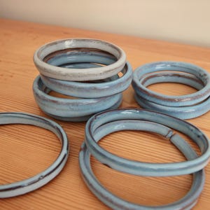 May include: A collection of seven circular ceramic rings with a smooth, glossy finish. The rings feature a light blue glaze with subtle variations in tone and dark brown edges along the rims. The items are arranged in small stacks and individually.