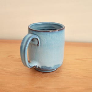 May include: A ceramic mug featuring a smooth, light blue glaze that transitions into a darker blue tone near the rim and base. The mug has a sturdy, curved handle on the side and a cylindrical body with a slightly flared rim, showcasing a glossy finish.