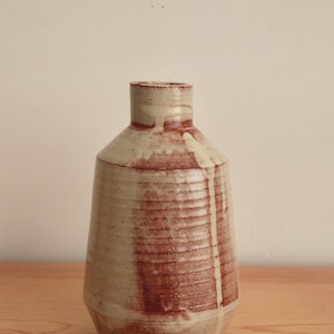 May include: A cylindrical ceramic vase with a narrow neck and a tapered base. The surface features a cream-colored glaze with vertical streaks of reddish-brown pigment and horizontal ridges. A small square stamp with the letters ep is visible.