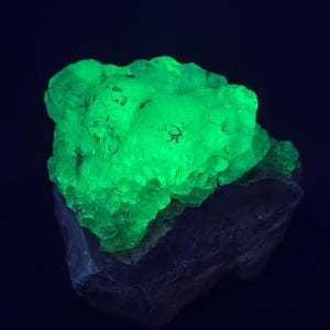 May include: A glowing green mineral specimen, possibly fluorite, sits atop a dark gray rock base. The green portion has a bubbly, textured surface, and the overall effect is striking against the dark background. The mineral appears to be illuminated from within.
