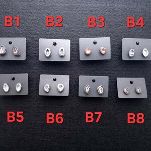 May include: A collection of eight pairs of stud earrings displayed on gray cards. The earrings feature various natural stone designs in shades of white, gray, brown, and gold. Each card is labeled with a red letter and number combination.