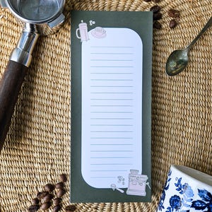May include: A green notepad with a white interior and lined pages, featuring coffee-themed illustrations. A coffee machine, cup, and saucer are depicted. A coffee maker handle, spoon, and a cup with a blue floral design are also visible.