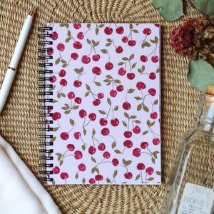 May include: A spiral-bound notebook with a light pink cover featuring a pattern of red cherries and green leaves. A white pen with gold accents rests beside the notebook on a woven mat. A clear glass bottle with a cork stopper is also visible.