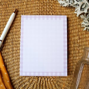 May include: A white pen with gold accents rests on a woven surface next to a notepad with a lavender gingham border. An orange scarf with white polka dots and a clear glass bottle are also visible.