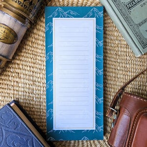 May include: A teal notepad with a white mountain design border and lined paper. The notepad is rectangular and sits on a woven mat, next to a brass telescope, a leather camera case, and a blue book.