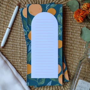 May include: A notepad with a teal background featuring orange fruit and green leaves. A white pen and a glass jar with a cork lid are on the woven surface. The notepad has a white arched top with lined paper.
