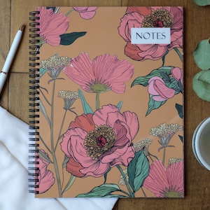 May include: Spiral-bound notebook with a floral design featuring pink and coral flowers on a tan background. The word "NOTES" is on a white label. A white pen with gold accents rests beside the notebook.