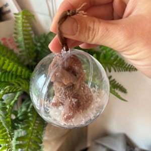 K&ouml;nnte beinhalten: Ein transparenter Glas-Anh&auml;nger mit einer braunen Hundefigur im Inneren. Der Anh&auml;nger ist mit wei&szlig;em Glitzer gef&uuml;llt und hat eine braune Schnur zum Aufh&auml;ngen.