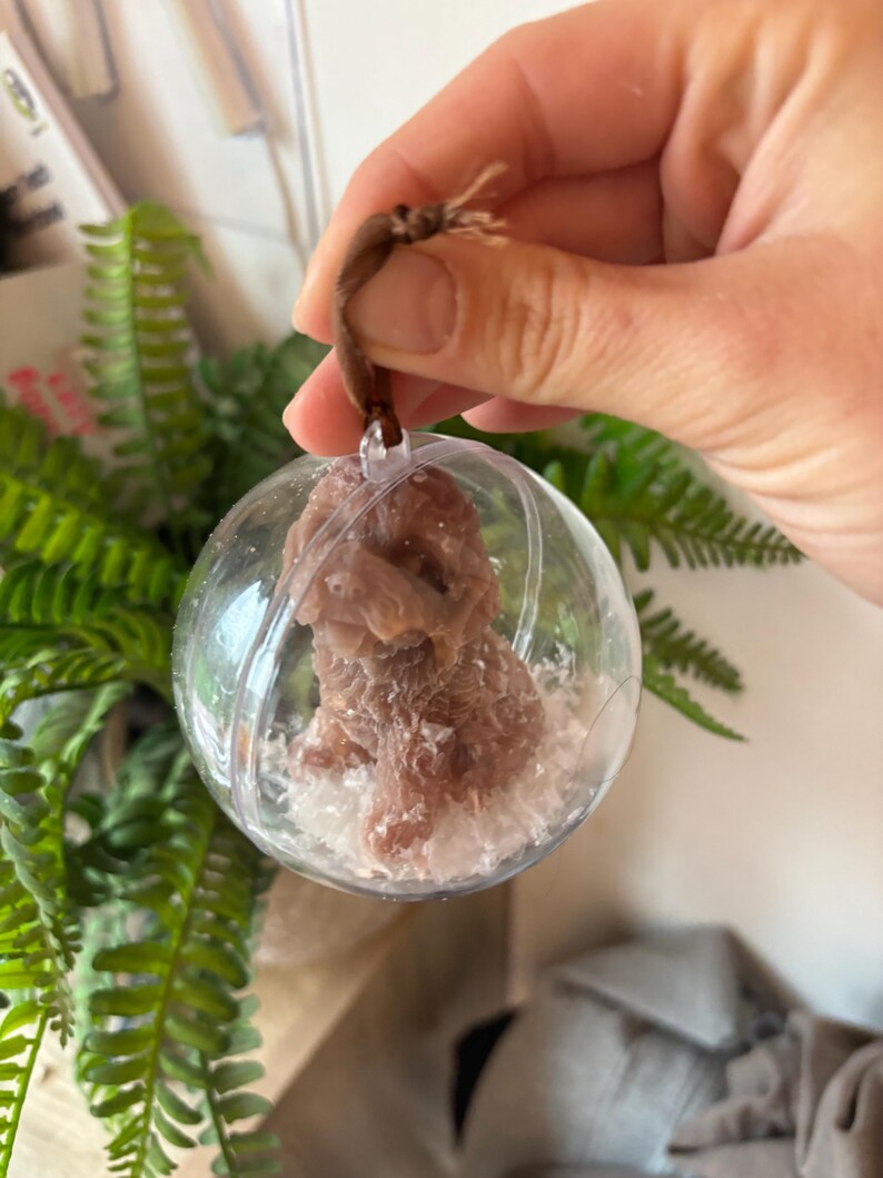 K&ouml;nnte beinhalten: Ein transparenter Glas-Anh&auml;nger mit einer braunen Hundefigur im Inneren. Der Anh&auml;nger ist mit wei&szlig;em Glitzer gef&uuml;llt und hat eine braune Schnur zum Aufh&auml;ngen.