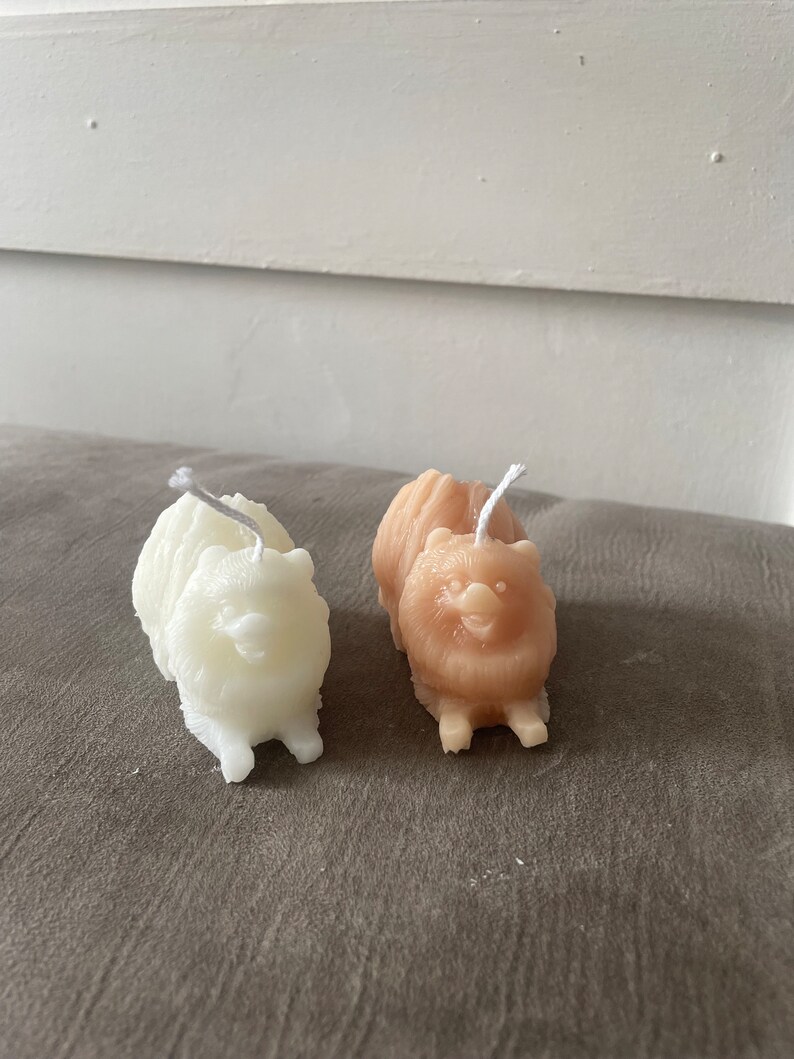 May include: Two small dog-shaped candles, one white and one light brown, with a wick sticking out of the top of each. The candles are sitting on a grey textured surface.