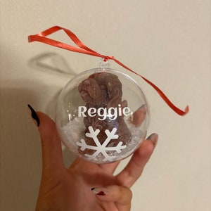 K&ouml;nnte beinhalten: Ein transparenter Weihnachtsbaumschmuck aus Glas mit einem braunen Pfotenabdruck im Inneren. Der Schmuck ist mit wei&szlig;em Kunst-Schnee und einer wei&szlig;en Schneeflocke verziert. Der Name "Reggie" ist auf dem Schmuckst&uuml;ck geschrieben.
