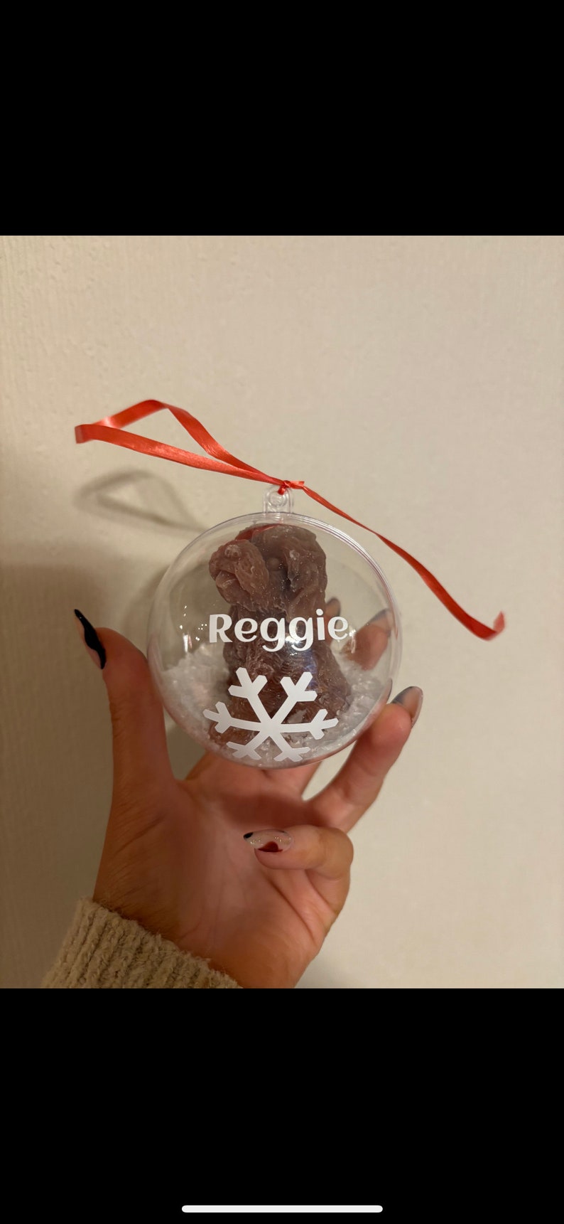 K&ouml;nnte beinhalten: Ein transparenter Weihnachtsbaumschmuck aus Glas mit einem braunen Pfotenabdruck im Inneren. Der Schmuck ist mit wei&szlig;em Kunst-Schnee und einer wei&szlig;en Schneeflocke verziert. Der Name "Reggie" ist auf dem Schmuckst&uuml;ck geschrieben.