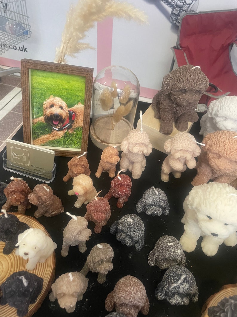 K&ouml;nnte beinhalten: Eine Pr&auml;sentation von hundef&ouml;rmigen Kerzen in verschiedenen Farben, darunter Braun, Wei&szlig; und Grau. Ein gerahmtes Foto eines Hundes und eine Glasglocke mit getrockneten Blumen sind ebenfalls vorhanden. Die Kerzen sind auf einer schwarzen Oberfl&auml;che angeordnet.