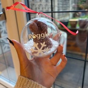 Puede incluir: Un adorno de Navidad de vidrio transparente con una huella de pata marrón en su interior. El adorno está lleno de purpurina blanca y tiene una cinta roja atada. El adorno está personalizado con el nombre "Reggie" y un copo de nieve.