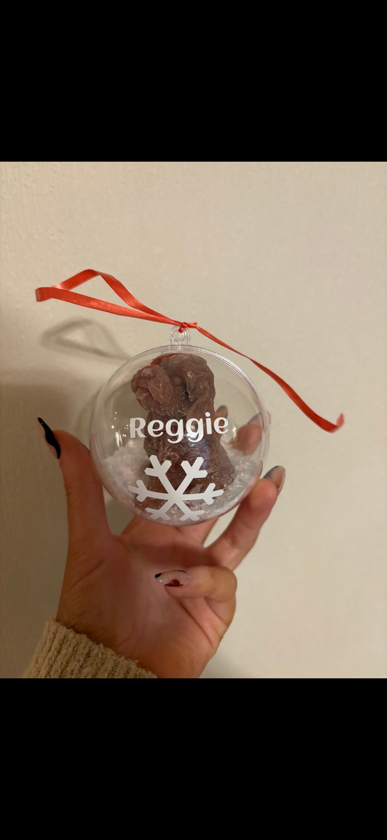 K&ouml;nnte beinhalten: Ein transparenter Weihnachtsbaumschmuck aus Glas mit einer braunen Hundefigur im Inneren. Der Schmuck ist mit einer wei&szlig;en Schneeflocke und dem Namen "Reggie" verziert.