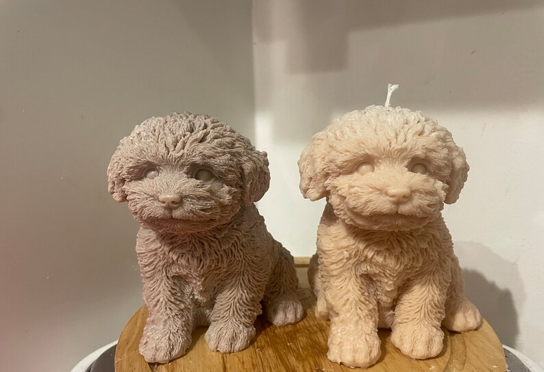 K&ouml;nnte beinhalten: Zwei hundef&ouml;rmige Kerzen in Beige- und Taupe-T&ouml;nen stehen nebeneinander. Die Kerzen sind mit strukturiertem Fell detailliert und haben eine sitzende Pose. Eine Kerze hat einen wei&szlig;en Docht. Die Kerzen stehen auf einer Holzoberfl&auml;che.