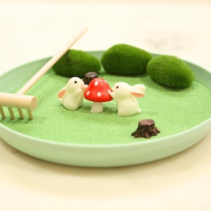 Puede incluir: Un jardín zen en miniatura en un plato verde claro. El jardín presenta una base de arena verde claro, pequeños conejos blancos, una seta roja, piedras musgosas verdes, un rastrillo de madera y un tocón de árbol. La escena está diseñada para crear un ambiente relajante.