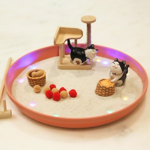 May include: A miniature zen garden in a coral-coloured circular tray. The garden features a wooden cat tree, two black and white cat figurines, small baskets, red and tan decorative balls, and a wooden rake.