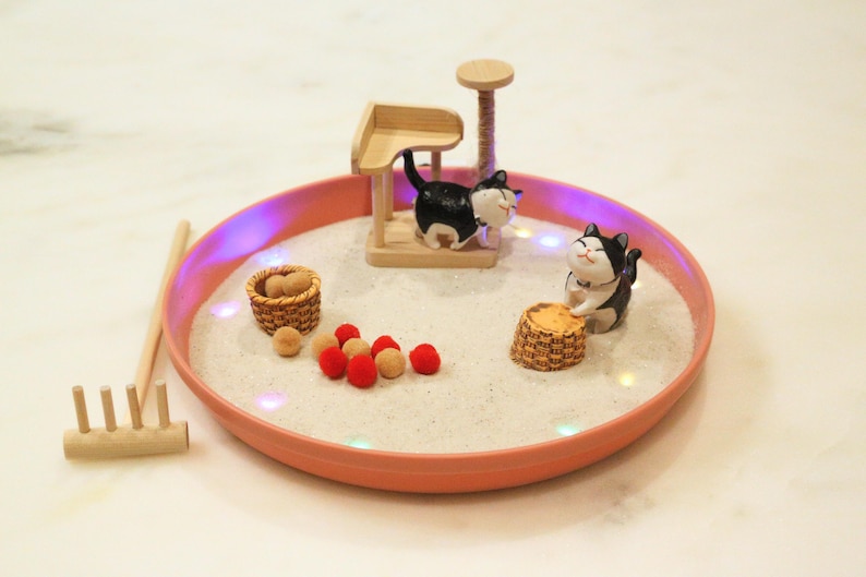 May include: A miniature zen garden in a coral-coloured circular tray. The garden features a wooden cat tree, two black and white cat figurines, small baskets, red and tan decorative balls, and a wooden rake.