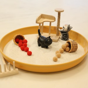 May include: A miniature zen garden in a yellow tray, featuring a black cat figurine, a cat scratching post, and two small baskets spilling red and tan spheres. A small wooden rake is also present.