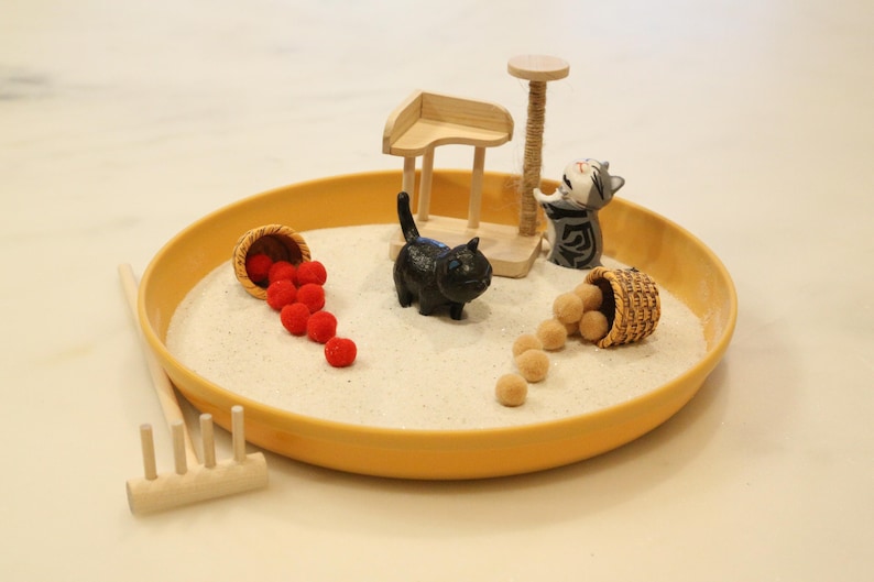 May include: A miniature zen garden in a yellow tray, featuring a black cat figurine, a cat scratching post, and two small baskets spilling red and tan spheres. A small wooden rake is also present.