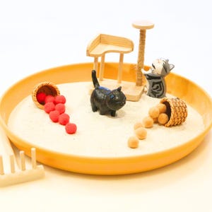 May include: A miniature cat-themed zen garden with a yellow tray filled with sand. It features a black cat figurine, a cat scratching post, and two small baskets spilling red and tan spheres. A wooden rake is also included.