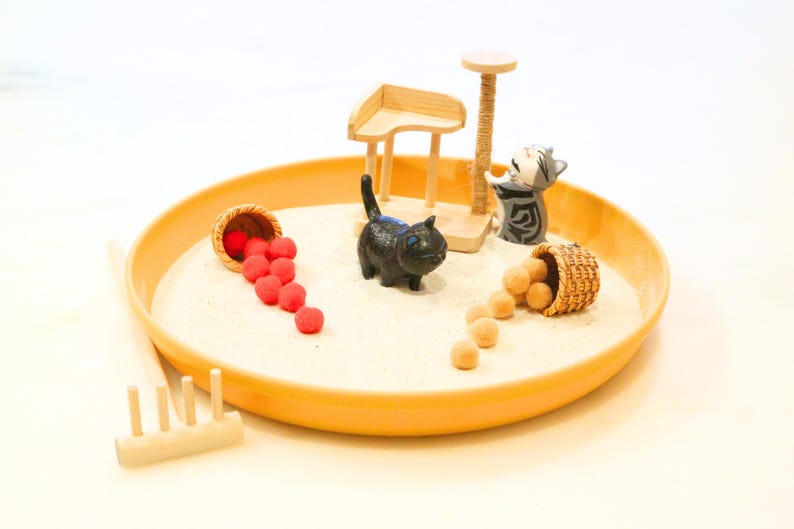May include: A miniature cat-themed zen garden with a yellow tray filled with sand. It features a black cat figurine, a cat scratching post, and two small baskets spilling red and tan spheres. A wooden rake is also included.
