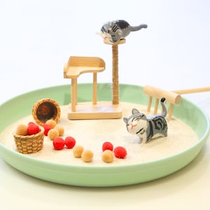 May include: A miniature zen garden scene with a light green tray filled with sand. It features small wooden furniture, a cat scratching post, and several cat figurines. Red and tan decorative balls and a small basket are also included.