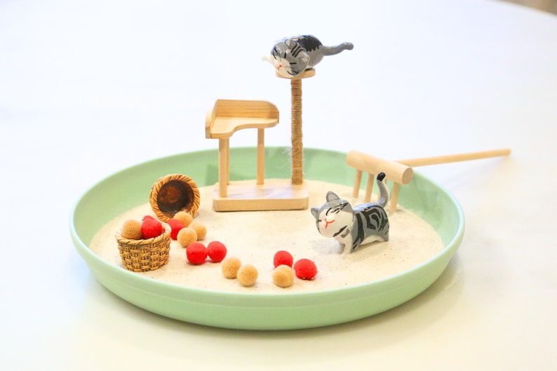 May include: A miniature zen garden scene with a light green tray filled with sand. It features small wooden furniture, a cat scratching post, and several cat figurines. Red and tan decorative balls and a small basket are also included.