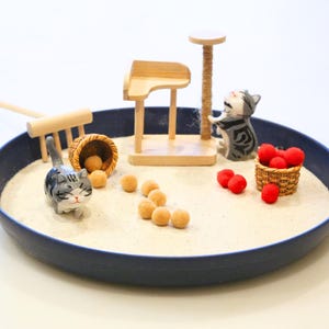 May include: A miniature zen garden scene in a navy blue bowl. It features a small wooden cat tree, a rake, two cat figurines, a basket of red pom-poms, and several tan-coloured spheres, all set on a bed of white sand.