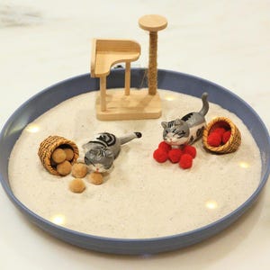 May include: A miniature cat-themed diorama in a blue dish. It features two grey and white cat figurines, small baskets with brown and red objects, and a wooden cat tree. The scene is set on a bed of white sand.