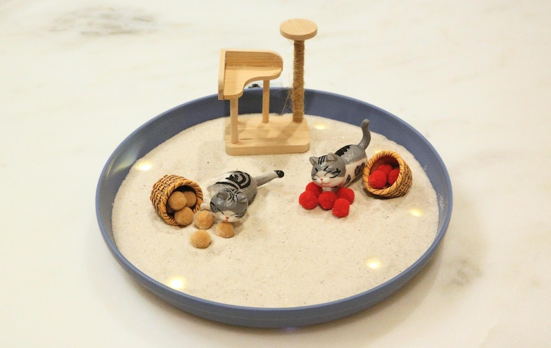 May include: A miniature cat-themed diorama in a blue dish. It features two grey and white cat figurines, small baskets with brown and red objects, and a wooden cat tree. The scene is set on a bed of white sand.