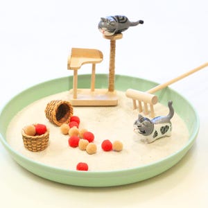 May include: A miniature zen garden with a light green tray filled with sand. It features a cat-themed design with cat figurines, a scratching post, a small rake, and decorative balls. A small basket is also included.